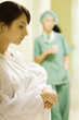 Photo: Young woman holding towel in hospital corridor, looking down, medical worker in background Young woman holding towel in hospital corridor, looking down, medical worker in background