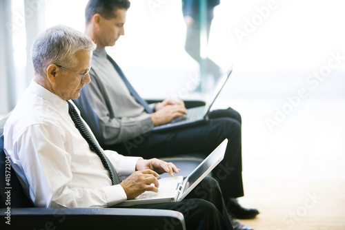 Foto: Two businessmen sitting in lobby, both using laptop computers, side view Two businessmen sitting in lobby, both using laptop computers, side view