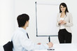 Woman standing in front of presentation board, smiling at camera, male colleague watching
