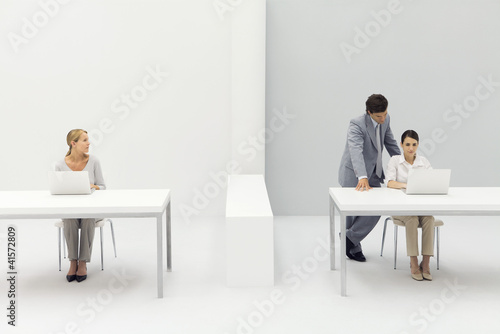Photo: Two women working in office, man standing behind one with his hand on her shoulder Two women working in office, man standing behind one with his hand on her shoulder