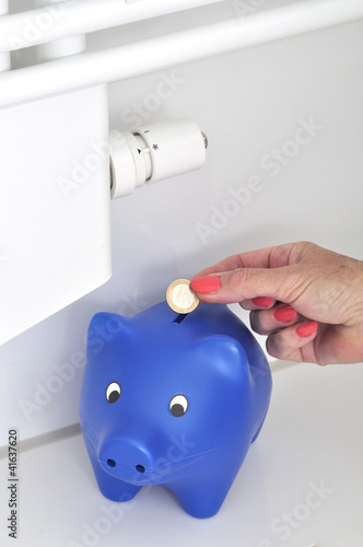 Foto: Frauenhand steckt 1 Euro in ein Sparschwein an einer Heizung Frauenhand steckt 1 Euro in ein Sparschwein an einer Heizung