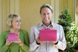 Foto: Mature couple holding pink gift boxes by Christmas tree, woman with small gift box looking at man, smiling Mature couple holding pink gift boxes by Christmas tree, woman with small gift box looking at man, smiling