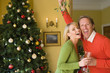 Couple by Christmas tree with champagne flutes, man holding up mistletoe, portrait