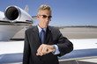 Businessman in sunglasses by aeroplane on runway, looking at wrist watch