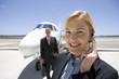 Photo: Businesswoman with hand on earpiece by businessman and aeroplane on runway, smiling, portrait Businesswoman with hand on earpiece by businessman and aeroplane on runway, smiling, portrait