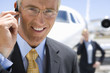 Photo: Businessman with hand on earpiece by businessman and aeroplane on runway, smiling, portrait Businessman with hand on earpiece by businessman and aeroplane on runway, smiling, portrait