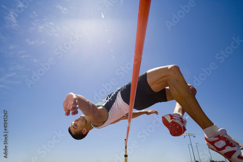 Photo: Young male athlete jumping over bar, low angle view (lens flare) Young male athlete jumping over bar, low angle view (lens flare)