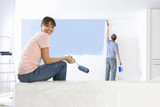 Foto: Woman holding paint roller and smiling as man paints living room wall in background Woman holding paint roller and smiling as man paints living room wall in background