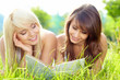 Two young beautiful smiling women reading book, sitting on grass