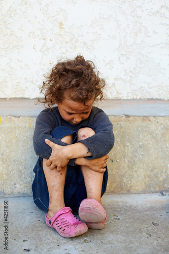 Photo: poor, sad little child girl sitting against the concrete wall poor, sad little child girl sitting against the concrete wall