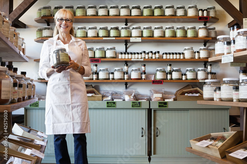 Photo: Portrait of a happy senior merchant standing with spice jar in store Portrait of a happy senior merchant standing with spice jar in store