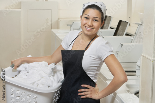 Photo: Portrait of a happy young female employee carrying laundry basket in Laundromat Portrait of a happy young female employee carrying laundry basket in Laundromat