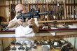 Foto: Mature merchant aiming with rifle in gun shop Mature merchant aiming with rifle in gun shop