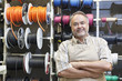 Photo: Portrait of a happy mature salesperson standing in front of electrical wire spool with arms crossed in hardware store Portrait of a happy mature salesperson standing in front of electrical wire spool with arms crossed in hardware store
