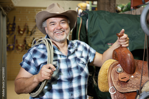 Foto: Happy mature cowboy with saddle and rope in feed store Happy mature cowboy with saddle and rope in feed store