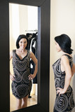 Foto: Happy mid adult woman wearing a stylish dress standing in front of mirror with hands on hips in boutique Happy mid adult woman wearing a stylish dress standing in front of mirror with hands on hips in boutique