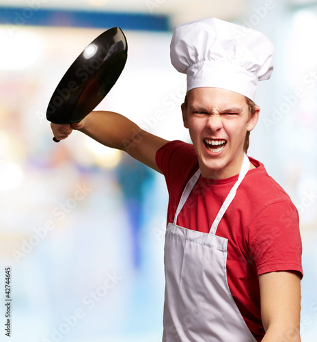 Photo: portrait of angry young cook man hitting with pan indoor portrait of angry young cook man hitting with pan indoor