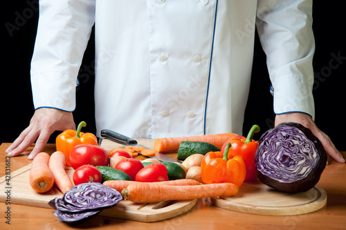 Photo: Chef standing by the kitchen table with variety of vegetables Chef standing by the kitchen table with variety of vegetables