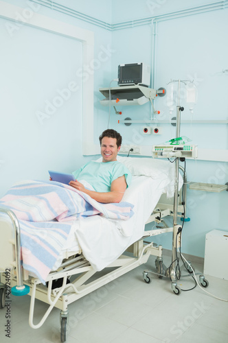 Photo: Joyful male patient holding a tactile tablet while lying on a bed Joyful male patient holding a tactile tablet while lying on a bed