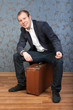 Foto: A young man sits on an old brown suitcase in the interior A young man sits on an old brown suitcase in the interior