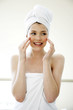 Foto: A woman with her hair and body wrapped up in towel smiling while applying under eye cream A woman with her hair and body wrapped up in towel smiling while applying under eye cream