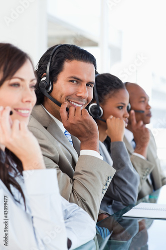 Photo: Young smiling call centre worker working among his business team Young smiling call centre worker working among his business team