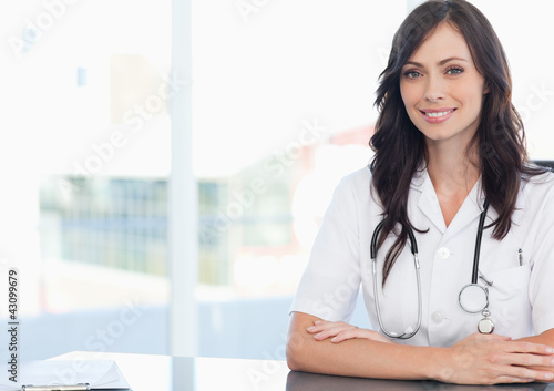 Photo: Young smiling doctor sitting at the desk near a clipboard Young smiling doctor sitting at the desk near a clipboard