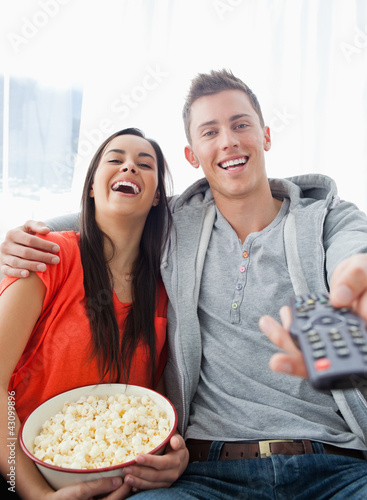Photo: A laughing couple sitting on the couch with popcorn as they watc A laughing couple sitting on the couch with popcorn as they watc