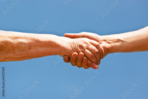 Photo: Handschlag einer alten Frau und einem jungen Mann vor blauem Him Handschlag einer alten Frau und einem jungen Mann vor blauem Him