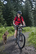 Woman riding mountain bike on wooded path with dog