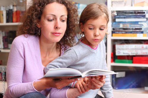 Photo: Daughter with her mother read book in room with bookshelves Daughter with her mother read book in room with bookshelves