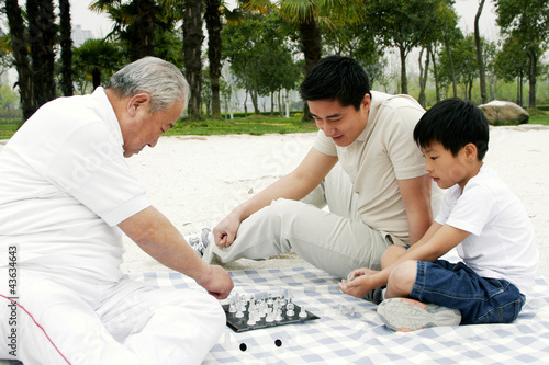 Photo: A boy playing chess with his grandfather on the lakeside while his father is watching A boy playing chess with his grandfather on the lakeside while his father is watching