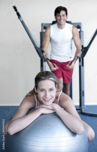 Photo: Woman resting on fitness ball, man exercising in the background Woman resting on fitness ball, man exercising in the background