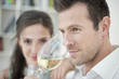 Man drinking white wine with his wife in the background