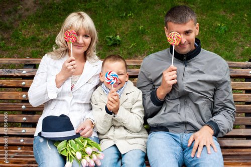 Photo: Funny family with toddler son having fun with candies on bench i Funny family with toddler son having fun with candies on bench i