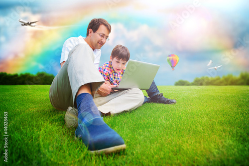 Photo: Adult man and his little son sitting on a grass at park and havi Adult man and his little son sitting on a grass at park and havi