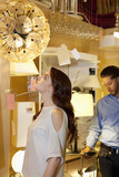 Photo: Beautiful young woman looking at lighting fixture hanging while man browsing in background Beautiful young woman looking at lighting fixture hanging while man browsing in background