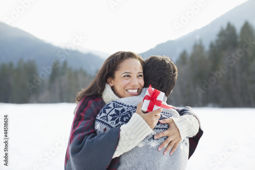 Photo: Smiling woman holding Christmas gift and hugging man in snow Smiling woman holding Christmas gift and hugging man in snow