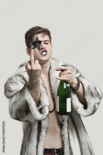 Photo: Portrait of young man with a rude gesture while holding beer bottle against gray background Portrait of young man with a rude gesture while holding beer bottle against gray background
