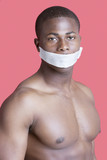 Photo: Portrait of shirtless African American man with tape over mouth against pink background Portrait of shirtless African American man with tape over mouth against pink background