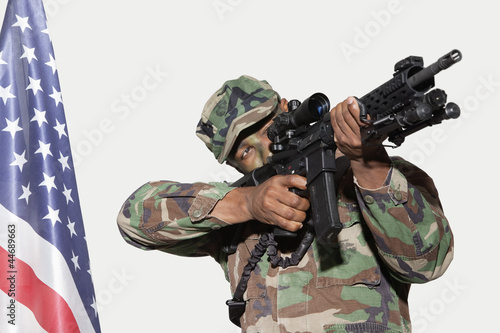 Photo: US Marine Corps soldier aiming M4 assault rifle with American flag against gray background US Marine Corps soldier aiming M4 assault rifle with American flag against gray background