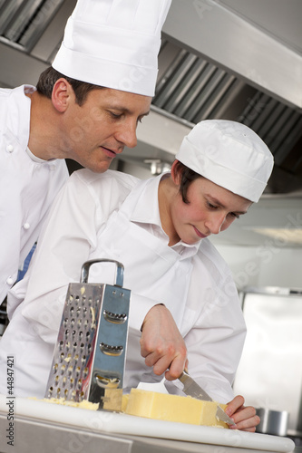 Photo: Chef watching trainee slice cheese in commercial kitchen Chef watching trainee slice cheese in commercial kitchen