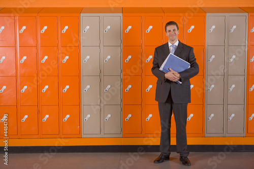 Photo: Teacher holding paperwork standing near school lockers Teacher holding paperwork standing near school lockers