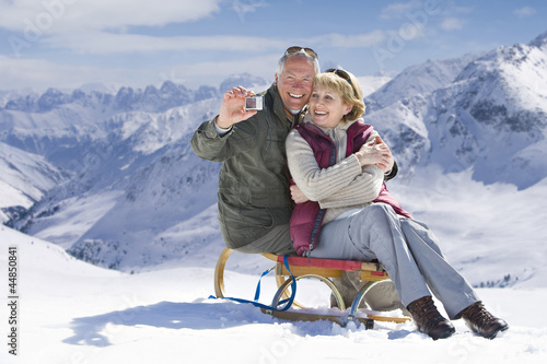 Photo: Senior couple on sled taking self-portrait with digital camera on snowy mountain Senior couple on sled taking self-portrait with digital camera on snowy mountain