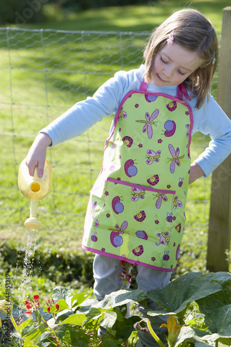 Photo: Girl in apron watering plants in garden with small watering can Girl in apron watering plants in garden with small watering can