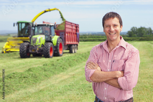 Photo: Portrait of smiling farmer with arms crossed in hay field with tractors Portrait of smiling farmer with arms crossed in hay field with tractors