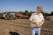 Foto: Farmer inspecting potatoes in sunny, rural field with tractors in background Farmer inspecting potatoes in sunny, rural field with tractors in background