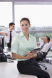 Photo: Portrait of smiling businesswoman leaning on desk and holding digital tablet in office with businessmen talking in background Portrait of smiling businesswoman leaning on desk and holding digital tablet in office with businessmen talking in background