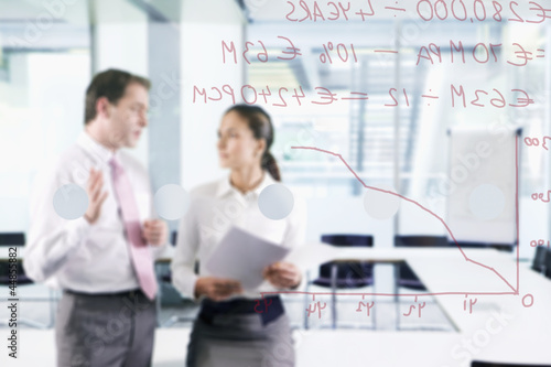 Photo: Businessman and businesswoman talking in conference room with graph on glass window in foreground Businessman and businesswoman talking in conference room with graph on glass window in foreground