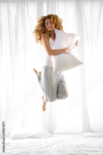 Photo: Happy blond woman holding pillow jumping on her bed Happy blond woman holding pillow jumping on her bed